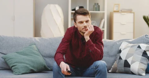 Young good looking man with a remote control sitting bored on the sofa, watching Stock Footage 87573988