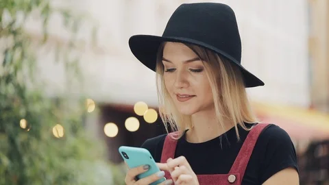 Young Good Looking Smiling Blond Girl Wearing a Black Hat Scrolling on her Stock Footage 122747400