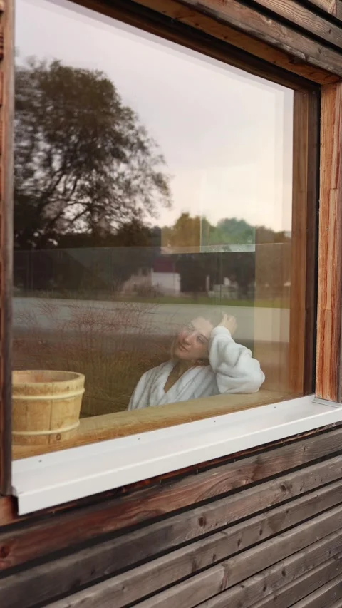 Young guest in white bathrobe rests inside wooden sauna seen through large windo Stock Footage 318219528
