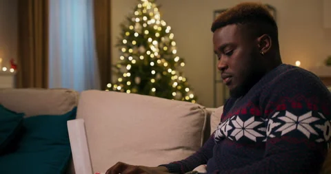 A young guy of African appearance sits on sofa in the living room and types on 스톡 동영상 211198408