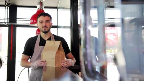 Young guy in apron holding paper package with take away meal and coffee inside Stock Footage 281248146