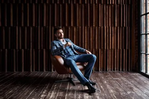 Young guy with a beard in a blue suit sits in a chair and looks into the dist Stock Photos