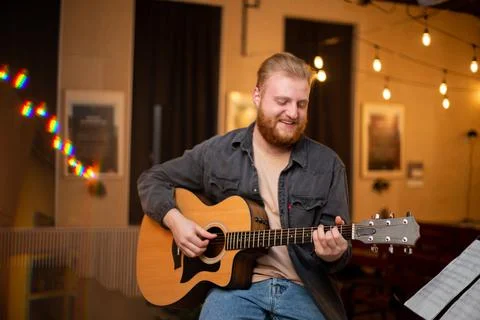 A young guy with a beard plays an acoustic guitar in a room with warm lighting Stock Photos