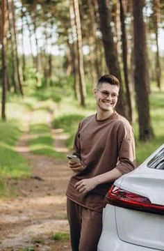 A young guy with a beard talking on the phone near his electric car. In his h Stock Photos
