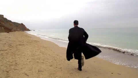 A young guy in a black coat runs along the beach against the backdrop of the sea Stock Footage 274694793