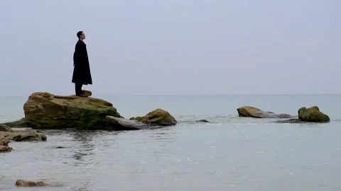 A young guy in a black coat is standing on the rocks in the sea Stock Footage 274695010