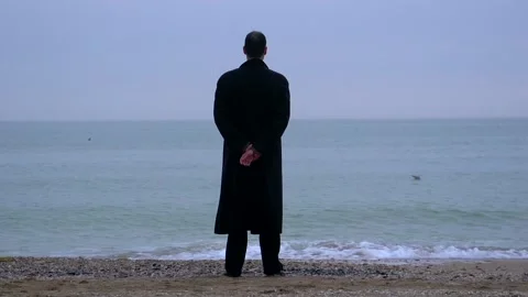 A young guy in a black coat stands against the background of the sky and the sea Stock Footage 274695046