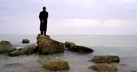A young guy in a black coat stands on the rocks in the sea Stock Footage 274875537