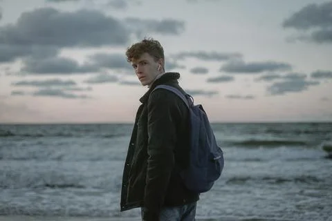 A young guy in a black denim jacket and curly hair stands on the seashore, lo Stock Photos