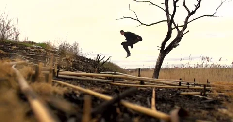 A young guy in a black long coat is jumping from a dry tree Stock Footage 274873820