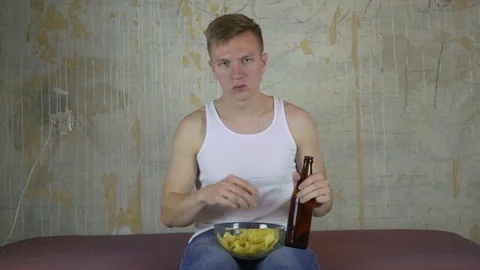 A young guy with a bottle of beer and chips watching TV 스톡 동영상 83023371