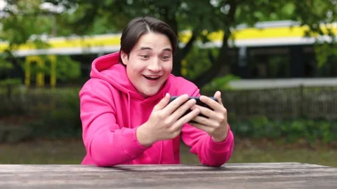 Young guy in bright clothes sits in park and watches funny videos on his phone. 스톡 동영상 253827530
