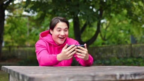 Young guy in bright clothes sits in park and watches funny videos on his phone. Vidéo 259490018