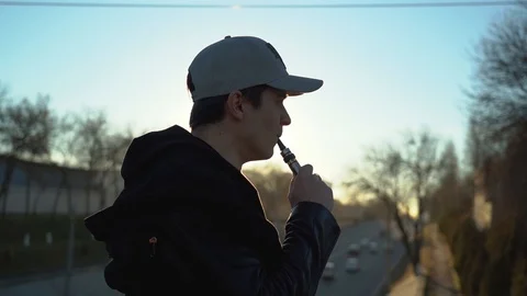 A young guy in a cap smokes an electronic cigarette at sunset, smoke burns in Stock Footage 99870840
