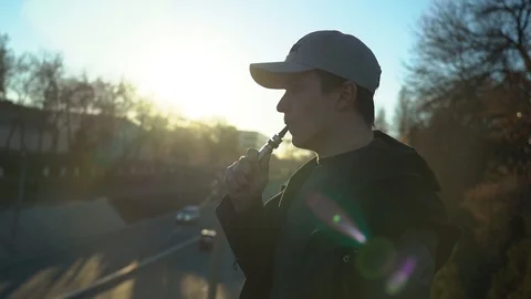 A young guy in a cap smokes an electronic cigarette at sunset, smoke burns in Stock Footage 99871163