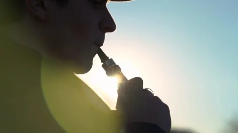 A young guy in a cap smokes an electronic cigarette at sunset, smoke burns in Stock Footage 99871869