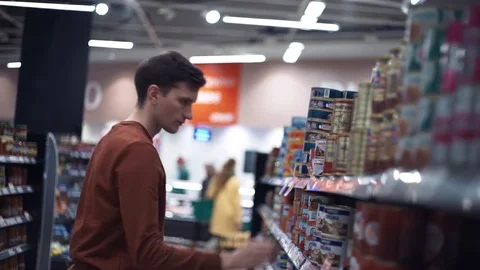 Young guy chooses canned pea on shelves full of food at supermarket Stock Footage 82990986