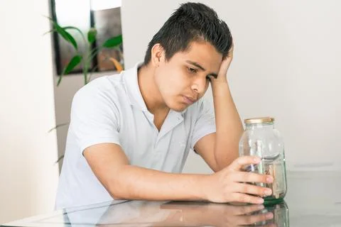 Young guy crestfallen looking at his savings in a glass jar. young man next t Stock Photos