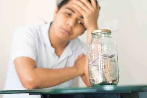Young guy crestfallen looking at his savings in a glass jar. young man next t Stock Photos