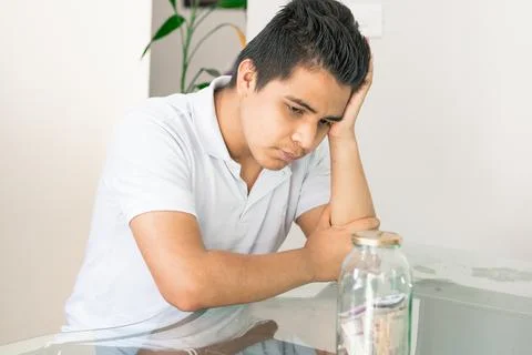 Young guy crestfallen looking at his savings in a glass jar. young man next t Stock Photos