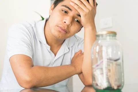 Young guy crestfallen looking at his savings in a glass jar. young man next t Stock Photos
