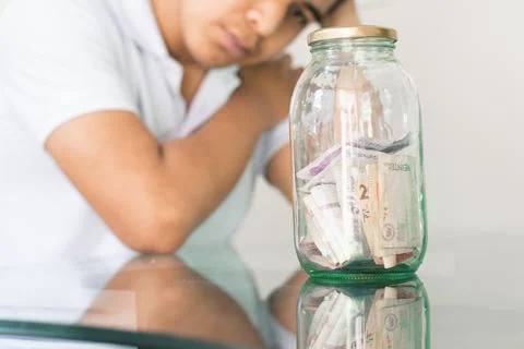 Young guy crestfallen looking at his savings in a glass jar. young man next t Stock Photos