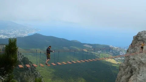 Young guy crosses abyss on rope bridge with insurance, experiencing adrenaline Stock Footage 80873895