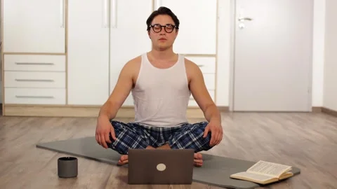 Young guy doing yoga at home with laptop end book.lazy yoga lessons on a karemat Vidéo 253827110