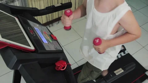 Young guy with dumbbells in his hands goes in for sports on a treadmill at home Stock Footage 200978723