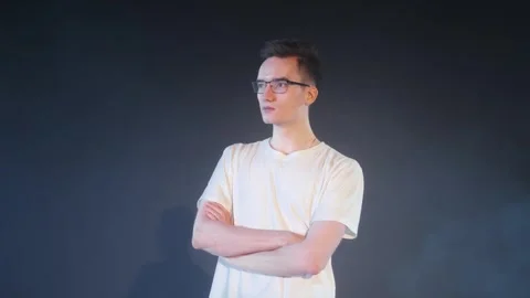 A young guy in glasses and a white T-shirt stands on a dark background. Stock Footage 299998159