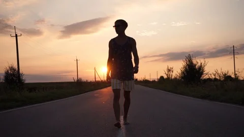 Young guy in glasses walks barefoot on asphalt. Incredible sunset Stock Footage 114631375