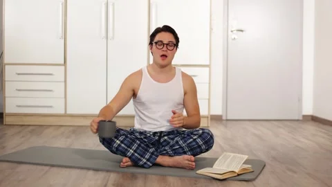 Young guy with glasses in white T-shirt drinks tea on carpet for sports at home Stock-Footage 253824718