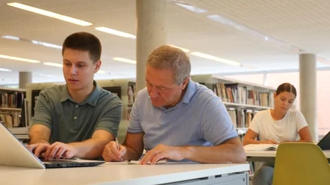 Young guy helping older man in laptop interface in public library Stock Footage 345514051