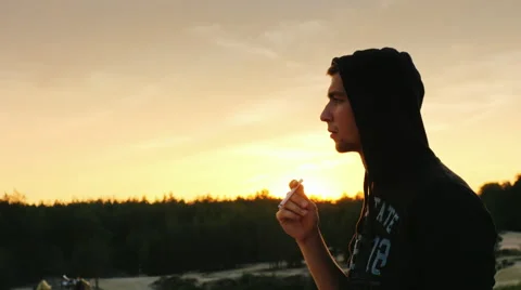 A young guy in a hood smoking a cigarette at sunset Stock Footage 66137598