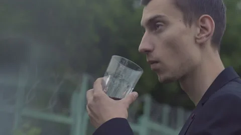 A young guy in a jacket stands and drinks whiskey from a glass Stock-Footage 155945856