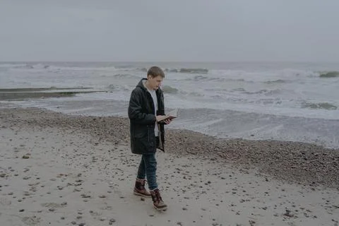 A young guy in a jacket walks along the seashore reading a book Stock Photos