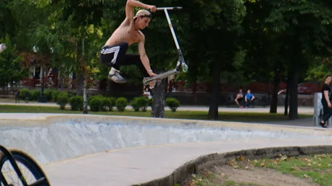 A young guy jumps and does tricks on a scooter in a skatepark Stock Footage 114246016