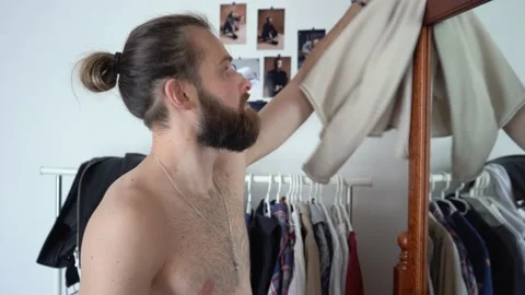 Young guy with long hair is looking into the mirror Putting together an outfit Stock Footage 147896643