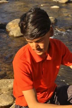 A young guy looking down and sitting on stone which beside of river Stock Photos