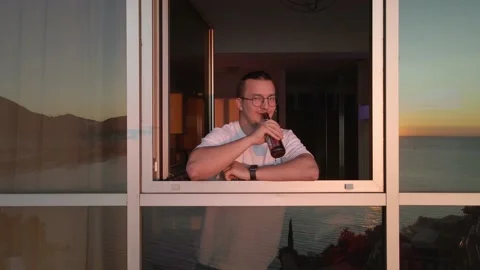 Young guy looking out of open window makes greeting gesture with bottle Video stock 159454698