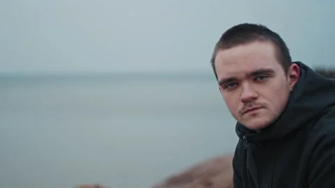 Young guy looks at the camera and sits on a stone, he is sad and cold Stock Footage 172266885