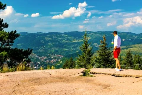 A young guy looks down at the valley and the mountains standing on a high poi Stock Photos