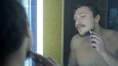 A young guy looks in the mirror and shaves his fair and curly beard Stock Footage 102673975