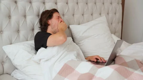 Young guy lying in the bed in the morning, yawn and start working on notebook Video stock 137975886