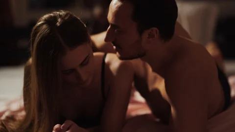 Young guy lying with pretty shy girl in bed in an intimate setting gently kisses Stock Footage 102731398