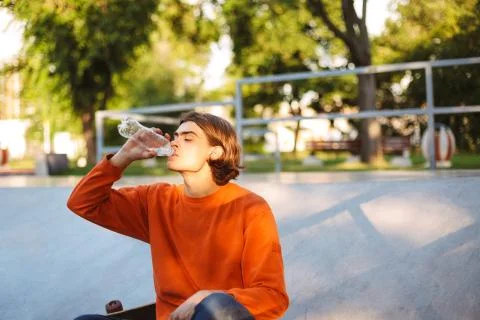 Young guy in orange pullover drinking water while spending time at skatepark Stock Photos