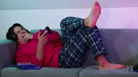 Young guy in pink sweater on sofa his legs crossed and playing game console Vidéo 253825610