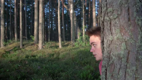 A young guy in a pink T-shirt looks out with fear from behind a tree Stock Footage 157306496
