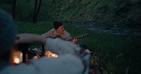 A young guy is playing in front of a fire near a mountain river, and his Stock Footage 114758967