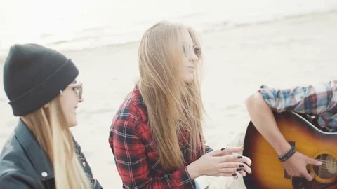 A young guy playing guitar sitting on a log, two attractive blond women clap Video stock 75690355